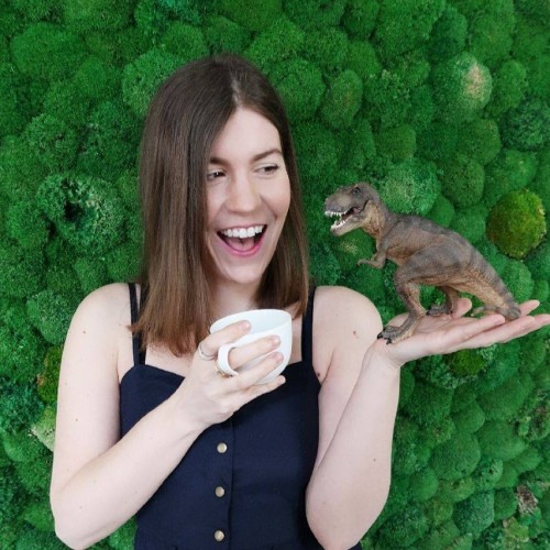 Portrait von Franziska Sattler mit Kaffeetasse und Plastikdino in der Hand vor einer moosbewachsenen Wand