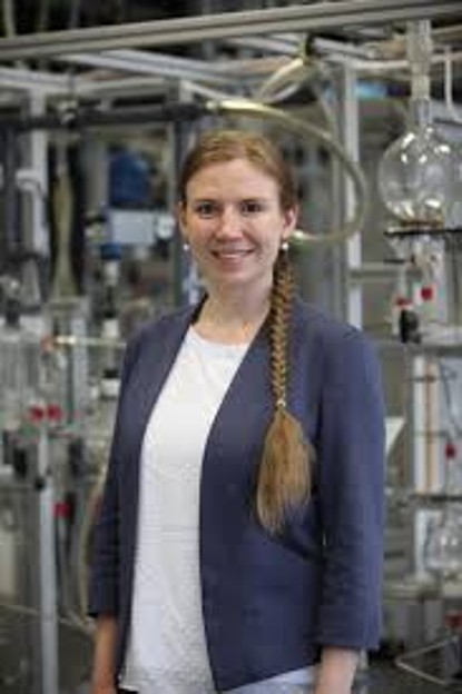 Bild von Kopf bis Hüfte von der lächelnden Dr. Kristina Zentel mit geflochtem über die Schulter geworfenem Zopf, in weißem Shirt und blauem Blazer, vor einer chemisch-technischen Anlage stehend